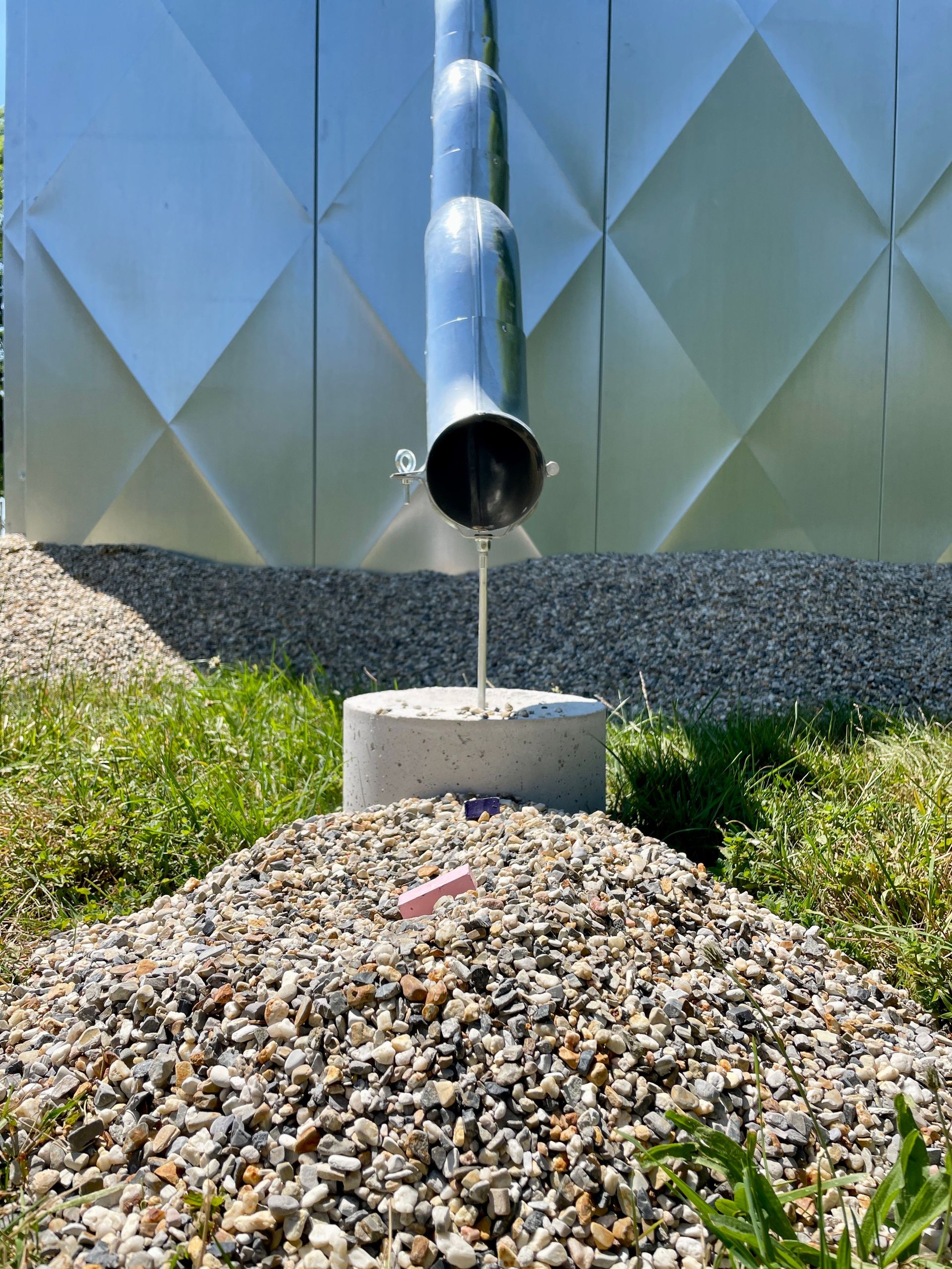 Stainless steel pipe releasing water onto gravel pile in a grassy outdoor setting.